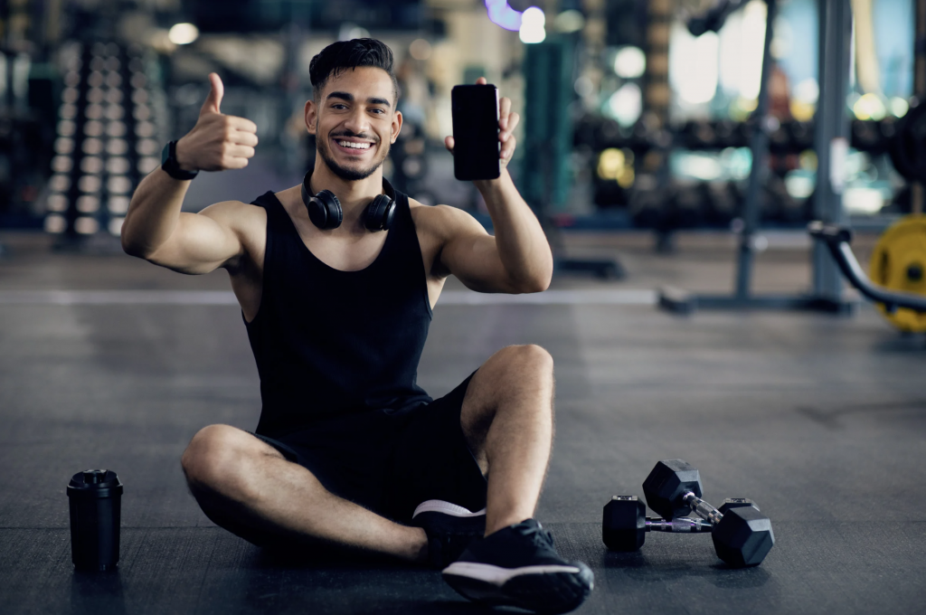 Gym member sitting on floor holding phone with thumbs up and dumbbells